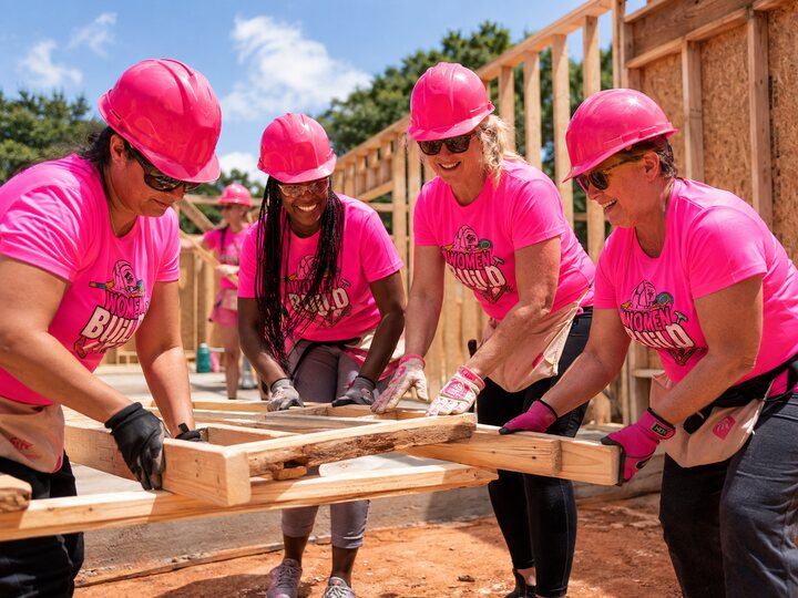 Women Build volunteers working together at a construction site in bright pink shirts and hard hats.